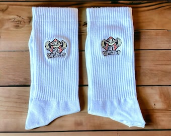 May include: Two white athletic socks with a ribbed texture. Each sock has an embroidered patch with a weightlifting design and the text "GOT MUSCLES". The socks are displayed on a wooden surface.