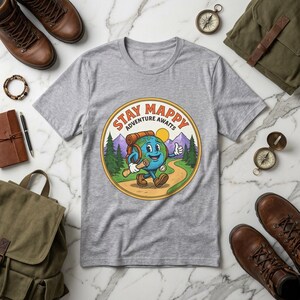以下が含まれることがあります： パスを歩く漫画の地球のキャラクターが描かれたグレーのTシャツ。バックパックを背負っています。「Stay Mappy Adventure Awaits」の文字が円形のエンブレムにデザインされています。ハイキングブーツ、コンパス、バックパックも表示されています。