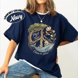 May include: Navy blue t-shirt with a graphic featuring a tree, whale, bee, and flowers. The text reads "Plant the Trees, Save the Bees, Clean the Seas." The shirt has a crew neck and short sleeves. The words "Comfort Colors" and "Navy" are in a circle on the upper left.