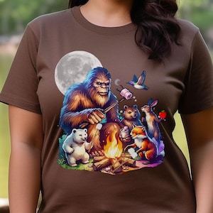 May include: A brown t-shirt with a graphic of a Bigfoot roasting marshmallows around a campfire with a polar bear, a fox, a rabbit, and a hummingbird. The moon is in the background.