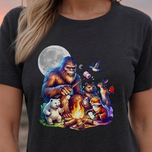 May include: A dark grey t-shirt with a graphic of Bigfoot roasting marshmallows around a campfire with a fox, rabbit, and two polar bears. The moon is in the background.