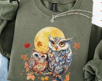 May include: An olive green sweatshirt featuring a detailed illustration of two owls perched on a branch with autumn leaves and a full moon. A silver necklace is draped across the sweatshirt. The sweatshirt is paired with blue jeans.