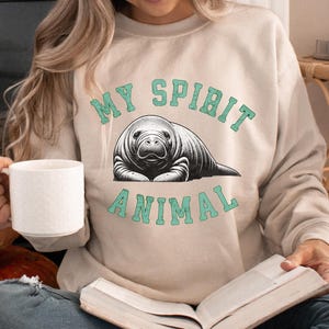 May include: A woman wearing a beige sweatshirt with the text "MY SPIRIT ANIMAL" in teal green lettering and a black and white illustration of a manatee. She is holding a white mug and a book.