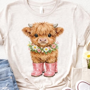 May include: A light beige t-shirt featuring a watercolor illustration of a Highland cow calf. The calf has brown fur, small horns, and is wearing a floral wreath and pink rain boots. The shirt is paired with denim shorts and white sneakers.