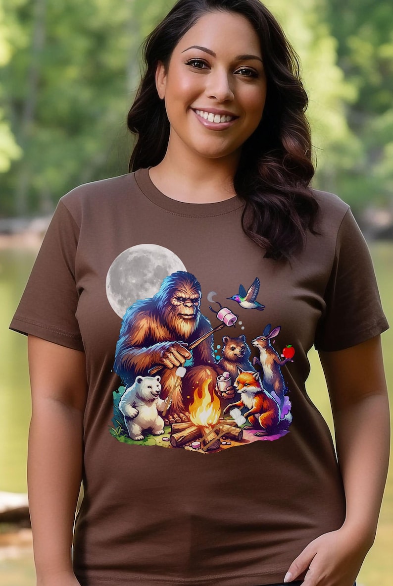 May include: A brown t-shirt with a graphic of a Bigfoot roasting marshmallows around a campfire with a polar bear, a fox, a rabbit, and a hummingbird. The moon is in the background.