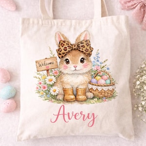 Pode incluir: Uma sacola creme com uma ilustra&ccedil;&atilde;o em aquarela de um coelho usando um la&ccedil;o e botas com estampa de leopardo. O coelho &eacute; cercado por flores e uma cesta de ovos de P&aacute;scoa. O nome "Avery" est&aacute; escrito em rosa.