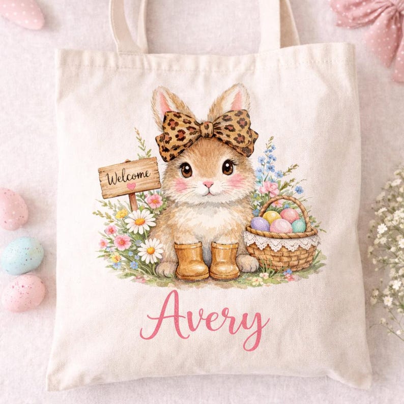 Pode incluir: Uma sacola creme com uma ilustra&ccedil;&atilde;o em aquarela de um coelho usando um la&ccedil;o e botas com estampa de leopardo. O coelho &eacute; cercado por flores e uma cesta de ovos de P&aacute;scoa. O nome "Avery" est&aacute; escrito em rosa.