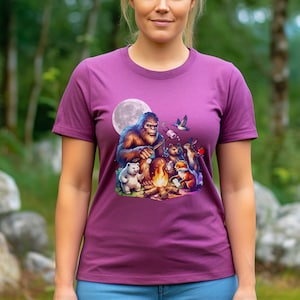 May include: A purple t-shirt with a graphic of Bigfoot roasting marshmallows around a campfire with a polar bear, a fox, a rabbit, and a bird. The moon is in the background.