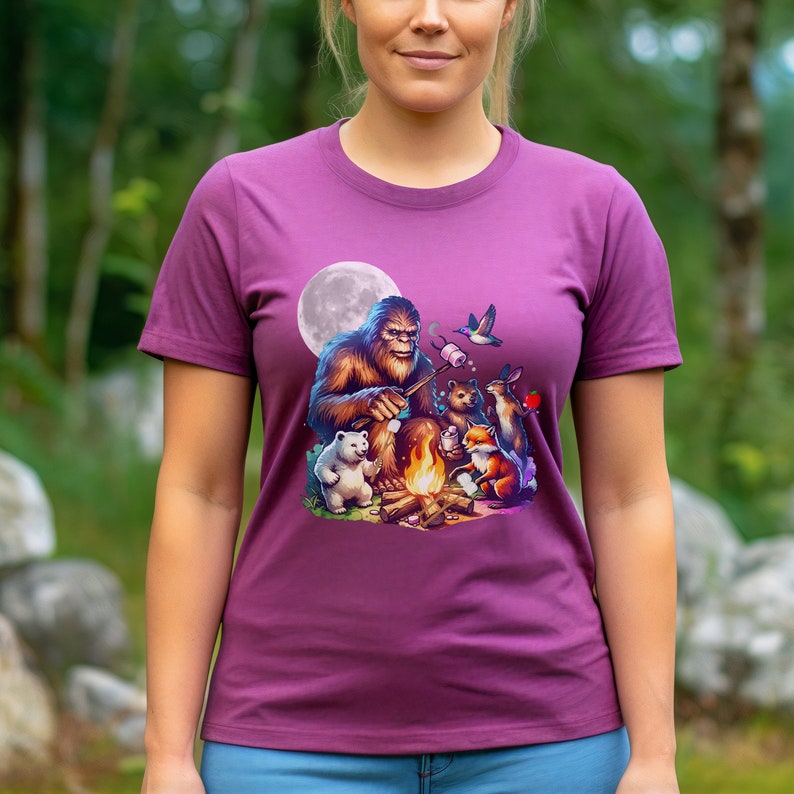 May include: A purple t-shirt with a graphic of Bigfoot roasting marshmallows around a campfire with a polar bear, a fox, a rabbit, and a bird. The moon is in the background.