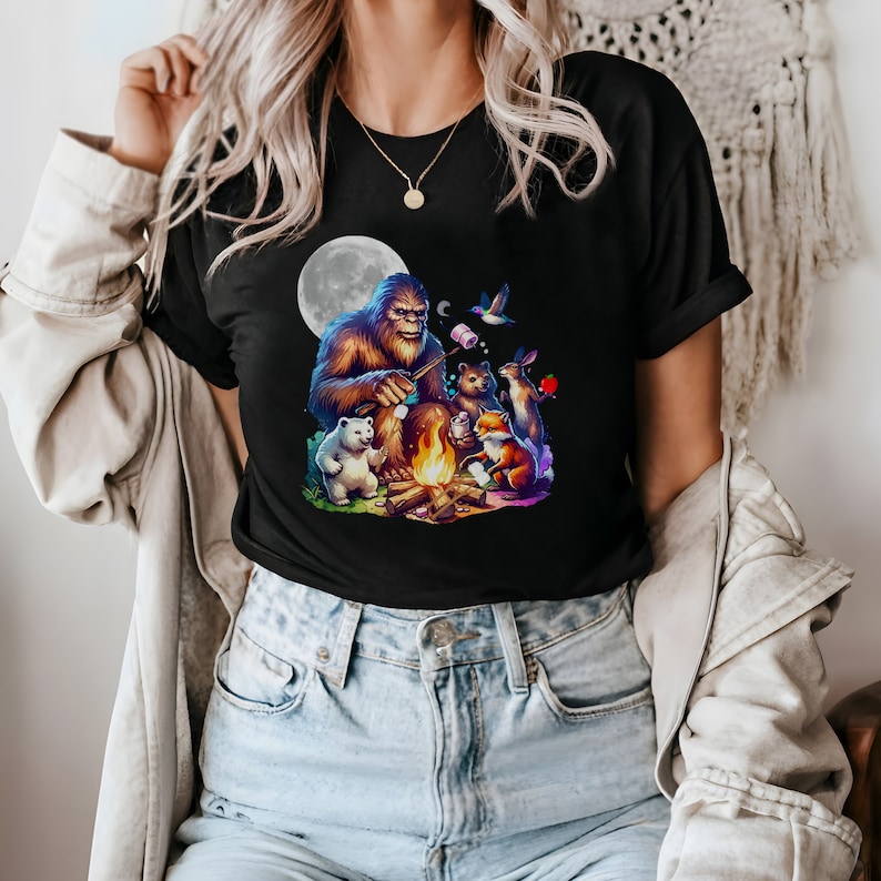 May include: A black t-shirt with a graphic of a Bigfoot roasting marshmallows around a campfire with a polar bear, a fox, a rabbit, and a bird. The graphic is set against a full moon.
