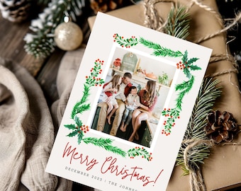 May include: A Christmas card with a watercolor border of green pine branches and red berries. The card features a photo of a family of four in a kitchen, with the text "Merry Christmas! December 2023 | The Johnson's" below the photo.