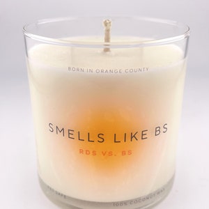 May include: A clear glass candle jar with a white candle inside. The candle has a label that reads "BORN IN ORANGE COUNTY" and "SMELLS LIKE BS RDS VS. BS". The bottom of the jar reads "PET SAFE" and "100% COCONUT WAX".