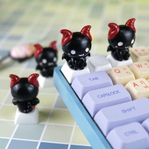 May include: Black and red devil character keycaps for a keyboard. The keycaps are on a blue keyboard with white and yellow keys. The keys have labels such as "Tab", "Capslock", and "Shift".