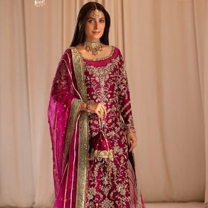 May include: A woman wearing a magenta embroidered dress with a matching sheer dupatta. The dress has gold and silver embellishments and a green border. The dupatta has a gold and silver border.