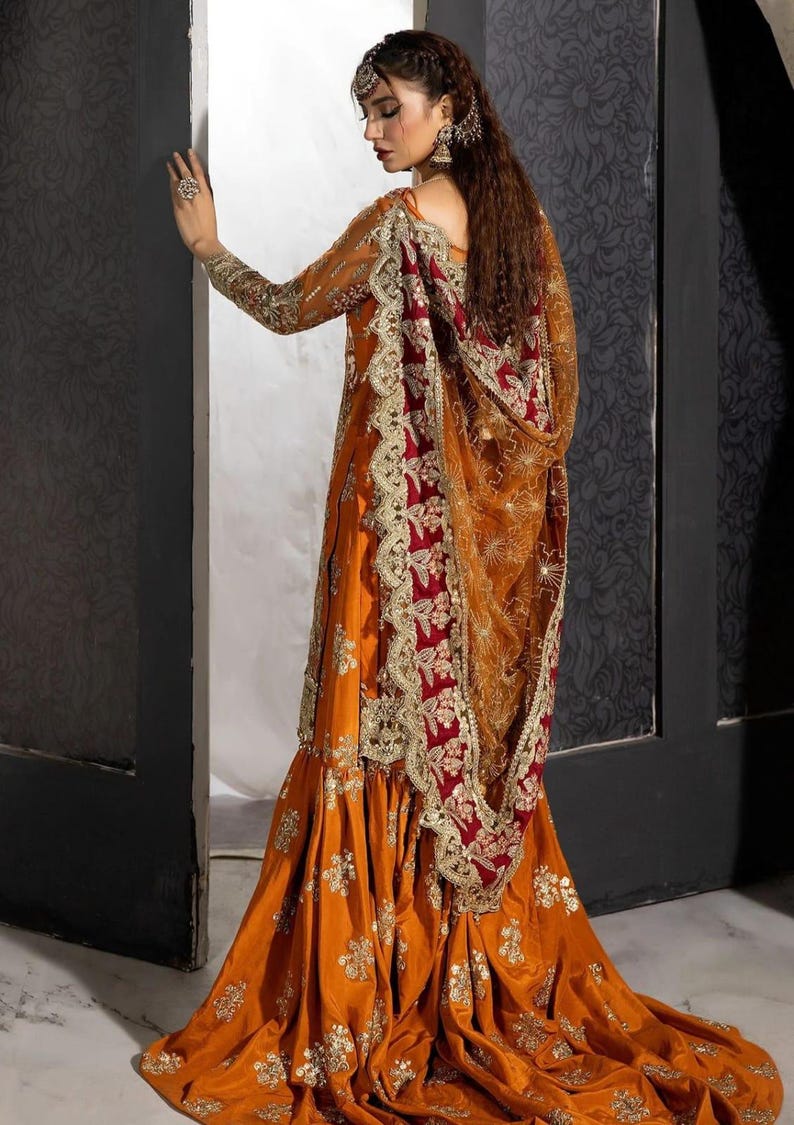 Op de afbeelding: Een vrouw draagt een lange, oranje en rode jurk met gouden borduurwerk. De jurk heeft een uitlopende rok en een doorschijnende dupatta met een rode rand.