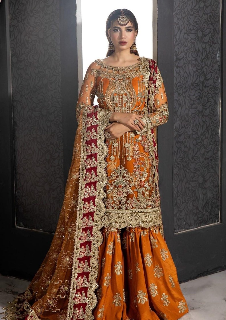 Op de afbeelding: Een vrouw draagt een lange, oranje en gouden geborduurde jurk met een maroon en gouden dupatta. De jurk heeft ingewikkelde bloemmotieven en gouden versieringen. De dupatta heeft een gegolfde rand met gouden en maroon borduurwerk.