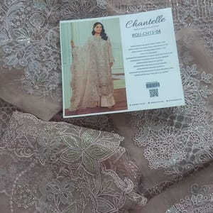May include: A light pink sheer fabric with silver embroidery and sequins. The fabric has a floral design and is decorated with small beads. The fabric is shown on a white background with a label that says "Chantelle Boutique Collection" and "BOU-CH13-04".