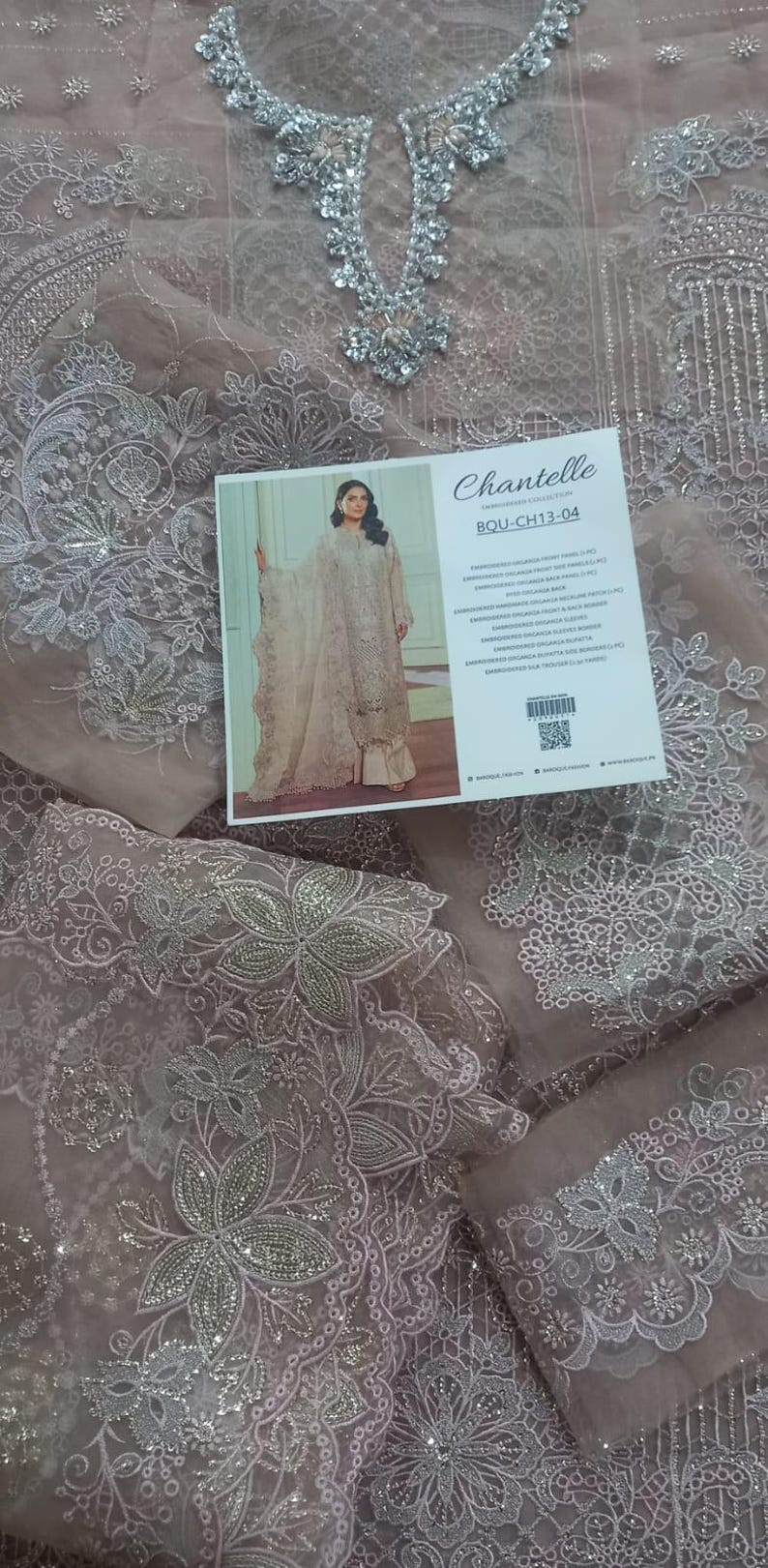 May include: A light pink sheer fabric with silver embroidery and sequins. The fabric has a floral design and is decorated with small beads. The fabric is shown on a white background with a label that says "Chantelle Boutique Collection" and "BOU-CH13-04".