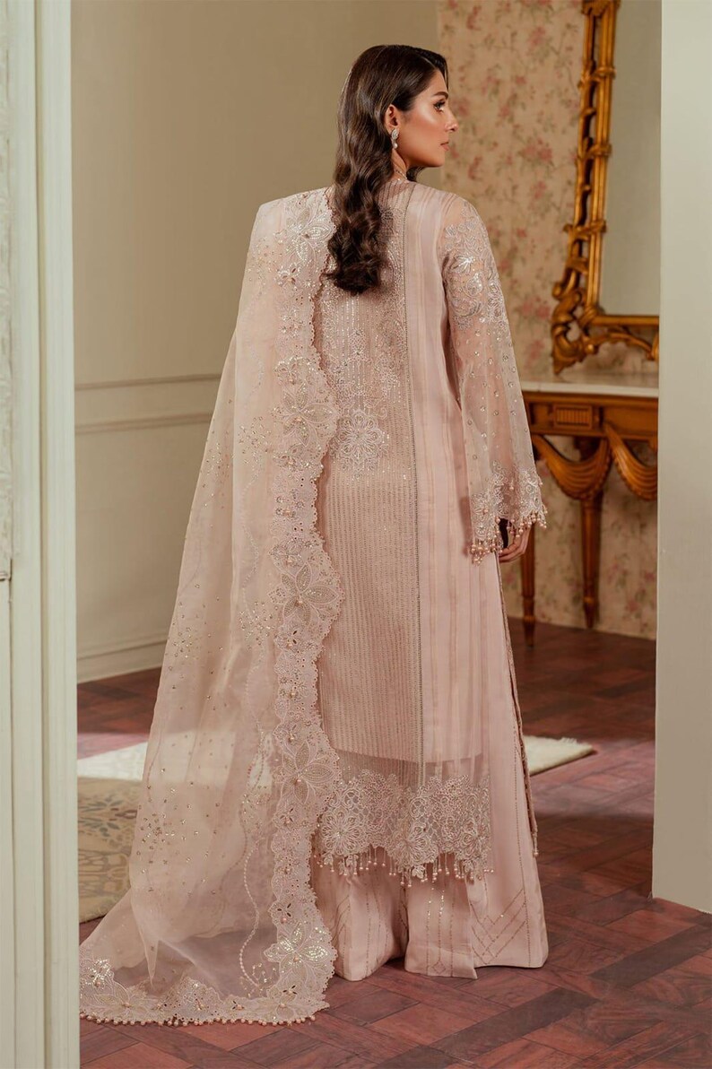 May include: A blush pink Pakistani shalwar kameez with intricate embroidery and lace detailing. The outfit features a long, flowing dupatta with a delicate floral pattern.