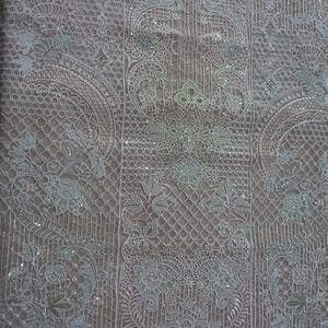 May include: A sheer, light brown fabric with a white embroidered design. The fabric is covered in small, sparkly sequins. The design features intricate floral patterns and geometric shapes. The fabric has a scalloped edge.