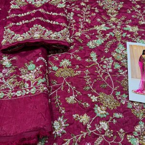 May include: A deep magenta fabric with intricate floral embroidery. The embroidery features delicate white and green flowers with gold accents. The fabric is embellished with small pearl-like beads.