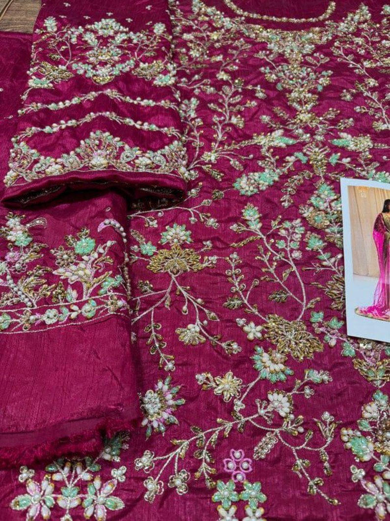 May include: A deep magenta fabric with intricate floral embroidery. The embroidery features delicate white and green flowers with gold accents. The fabric is embellished with small pearl-like beads.
