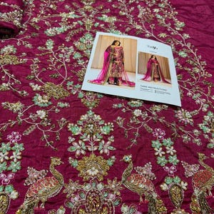 May include: A close-up of a magenta fabric with intricate embroidery featuring peacocks, flowers, and beads. The fabric is likely for a garment, such as a dress or a skirt.