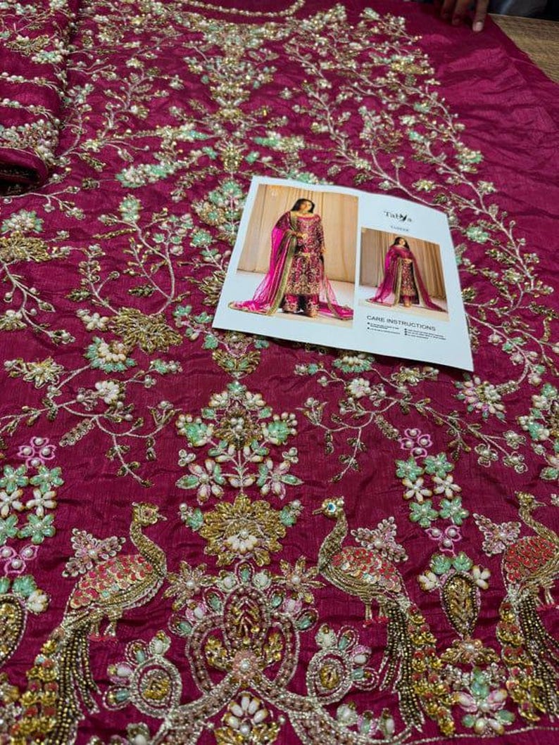 May include: A close-up of a magenta fabric with intricate embroidery featuring peacocks, flowers, and beads. The fabric is likely for a garment, such as a dress or a skirt.