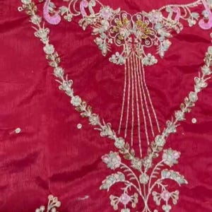 May include: A close-up of a red fabric with intricate embroidery in white, gold, and pink. The embroidery features floral designs and a central motif that resembles a stylised tree.