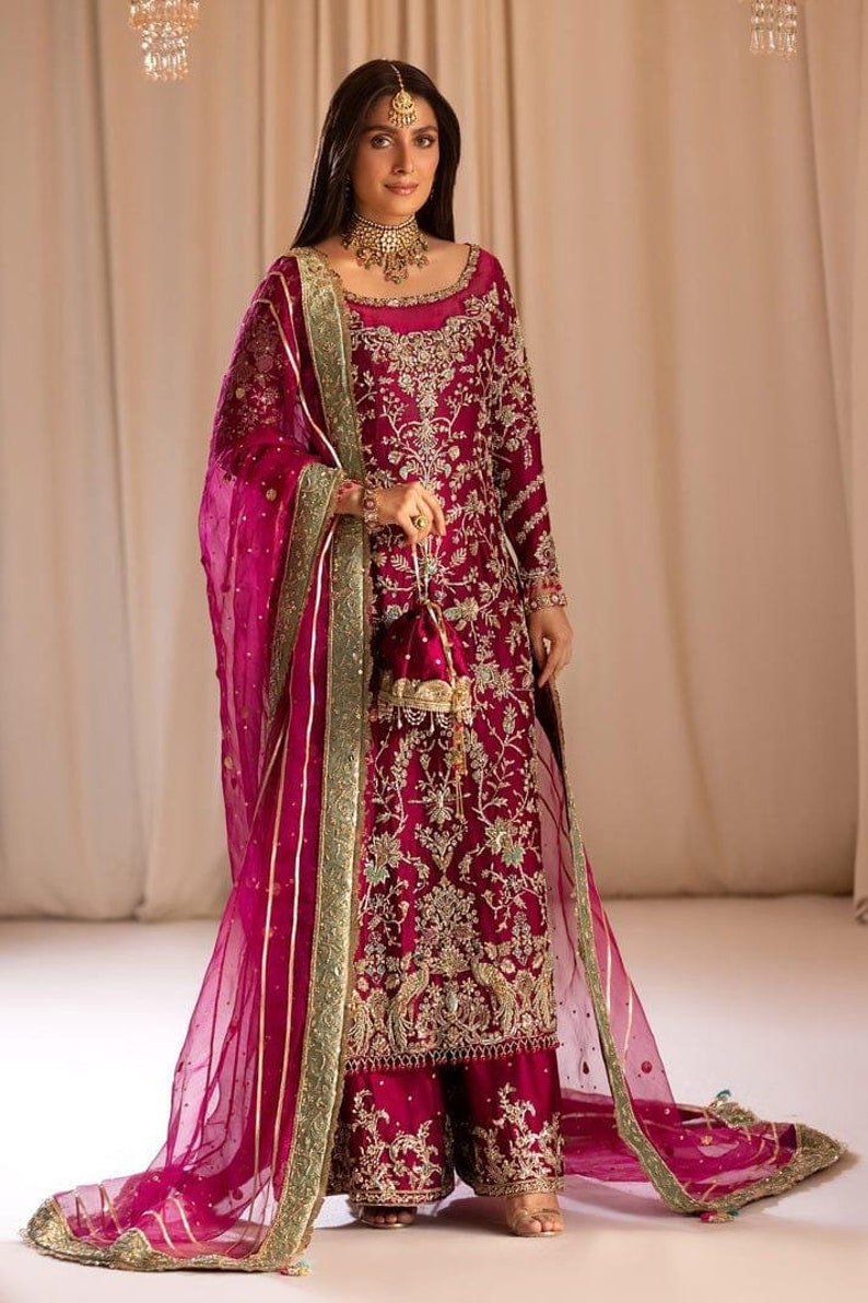May include: A woman wearing a magenta embroidered dress with a matching sheer dupatta. The dress has gold and silver embellishments and a green border. The dupatta has a gold and silver border.