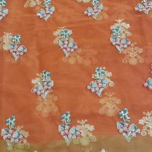 Op de afbeelding: Oranje doorzichtig stof met zilveren geborduurde bloemenmotieven. De stof heeft een delicate, kant-achtige rand.