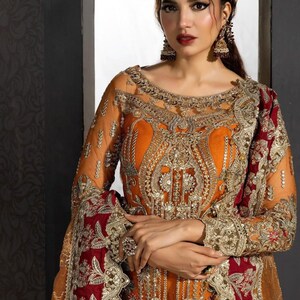 Op de afbeelding: Een vrouw die een lange, oranje en rode jurk draagt met ingewikkelde gouden borduursels en versieringen. De jurk heeft een doorschijnende overlay en een diepe halslijn. De vrouw draagt een traditionele Indiase hoofdtooi en oorbellen.