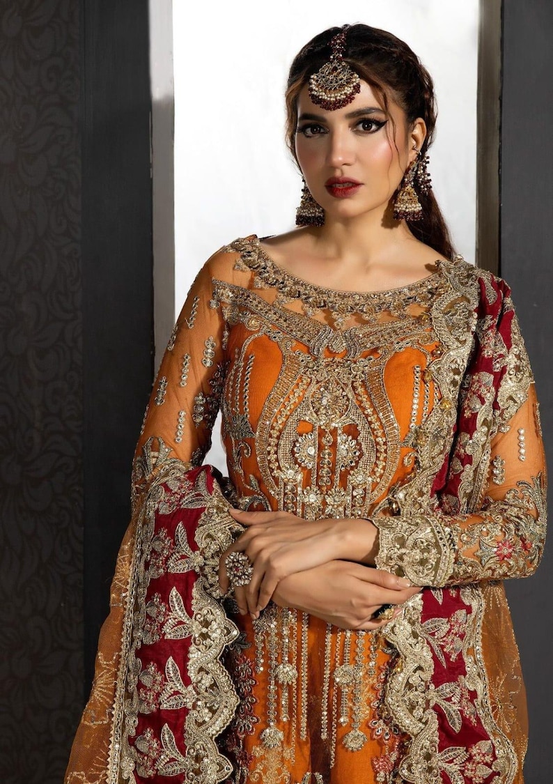 Op de afbeelding: Een vrouw die een lange, oranje en rode jurk draagt met ingewikkelde gouden borduursels en versieringen. De jurk heeft een doorschijnende overlay en een diepe halslijn. De vrouw draagt een traditionele Indiase hoofdtooi en oorbellen.