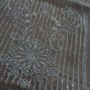May include: White lace fabric with silver sequins and beads. The fabric has a floral pattern and is lined with vertical rows of sequins.