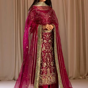 May include: A woman wearing a maroon and gold embroidered outfit with a matching sheer dupatta. The outfit features intricate floral and paisley designs. The dupatta has a gold border and is draped over the woman's shoulders.
