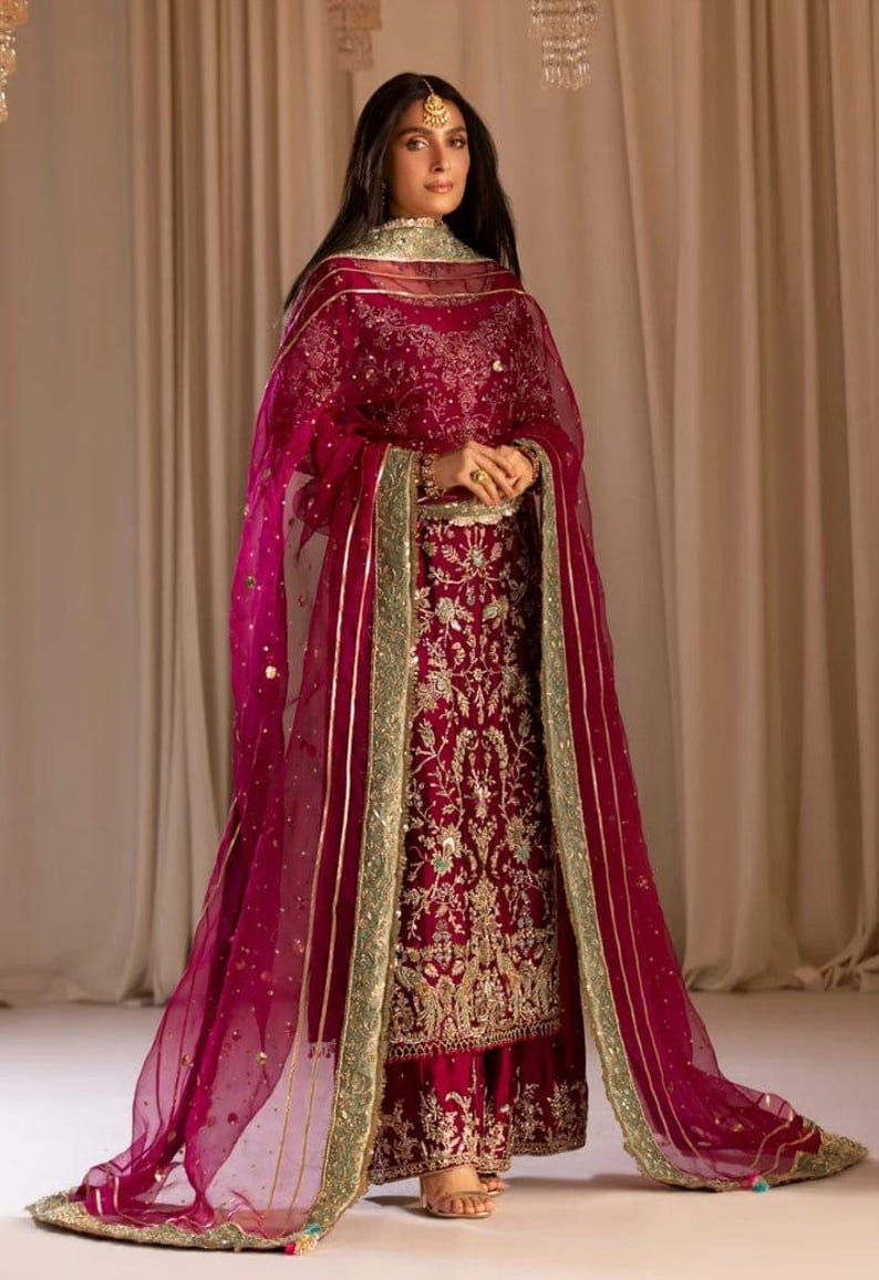 May include: A woman wearing a maroon and gold embroidered outfit with a matching sheer dupatta. The outfit features intricate floral and paisley designs. The dupatta has a gold border and is draped over the woman's shoulders.
