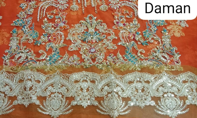Op de afbeelding: Oranje stof met ingewikkelde zilveren en turquoise borduurwerk. De stof heeft een delicate kantversiering. Het woord "Daman" staat in witte tekst op de stof.