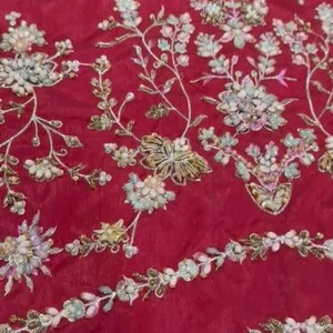May include: Red fabric with white and gold floral embroidery. The embroidery features delicate flowers and leaves with pearl-like embellishments. The fabric has a subtle sheen and a soft texture.