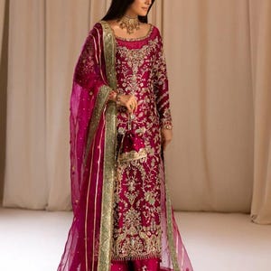 May include: A woman wearing a magenta embroidered dress with a matching sheer dupatta. The dress features gold and silver embellishments and a green border. The dupatta has a gold border and is draped over her shoulders.