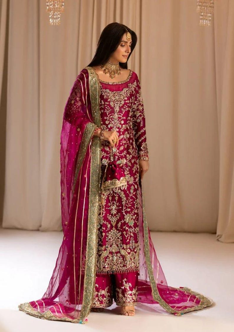 May include: A woman wearing a magenta embroidered dress with a matching sheer dupatta. The dress features gold and silver embellishments and a green border. The dupatta has a gold border and is draped over her shoulders.
