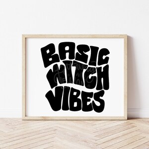 Pode incluir: Impressão em preto e branco com o texto "Basic Witch Vibes" em um estilo de fonte retrô e ondulado. A impressão é emoldurada em uma moldura de madeira marrom clara.