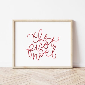 May include: A framed print with the words "the first noel" in red script on a white background. The frame is a light wood color, and the print is displayed against a white wall and wooden floor.