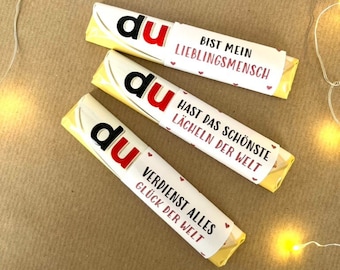 May include: Three yellow chocolate bars with white labels. The labels have the word "du" in red and a message in black. The messages are "Bist mein Lieblingsmensch", "Hast das schönste Lächeln der Welt", and "Verdienst alles Glück der Welt".