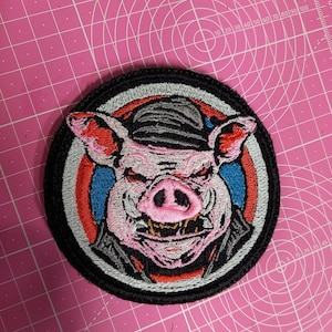 Puede incluir: Parche bordado con un cerdo rosa que lleva una chaqueta de cuero negra y un sombrero negro y blanco. El cerdo tiene una expresión amenazante con dientes afilados y un hocico rosa. El parche está rodeado por un círculo blanco, azul y rojo.