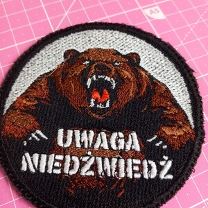 May include: A black and white embroidered patch with a brown bear roaring. The bear is surrounded by a white circle with the text "UWAGA NIEDZWIEDZ" in white.