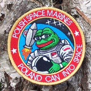 May include: A red, white and gold embroidered patch featuring a green cartoon frog wearing a space suit and holding a rocket. The patch reads "Polish Space Marines Poland Can Into Space".