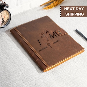 May include: Brown leather journal with personalized initials "J" and "M" with the names "Jackson" and "Marry" engraved on the front. The journal has a decorative flower design and a flap closure with two metal snaps.