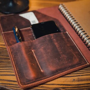 K&ouml;nnte beinhalten: Ein braunes Leder-Notizbuchcover mit einem Stifthalter, einer Tasche f&uuml;r das Telefon und einem spiralgebundenen Notizbuch. Das Cover hat einen Used-Look und ist mit wei&szlig;em Garn vern&auml;ht.