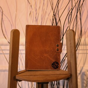 K&ouml;nnte beinhalten: Ein braunes Lederjournal mit einem personalisierten Monogramm auf der Vorderseite. Das Journal liegt auf einem Holzstuhl.