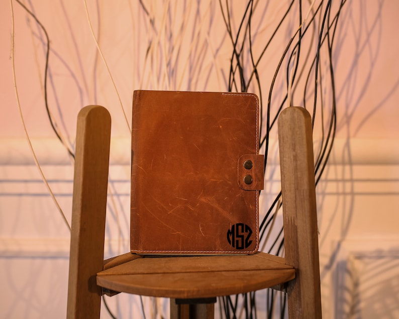 K&ouml;nnte beinhalten: Ein braunes Lederjournal mit einem personalisierten Monogramm auf der Vorderseite. Das Journal liegt auf einem Holzstuhl.